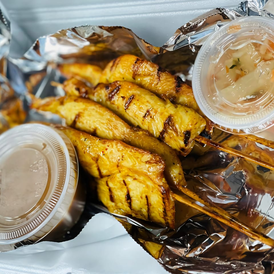 Grilled chicken satay skewers with peanut sauce and cucumber relish in a takeout container.
